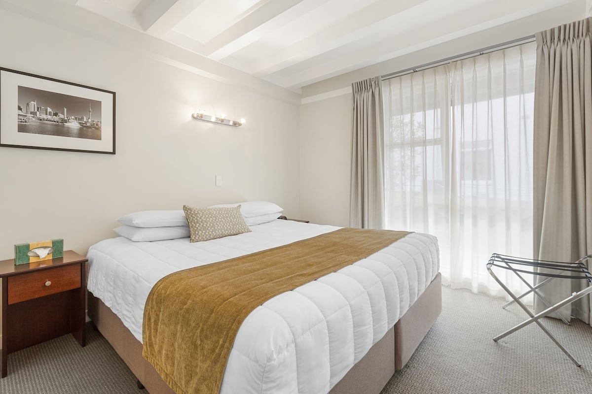 A hotel room with a large bed, a nightstand with a lamp, a framed picture on the wall, and a window with curtains.
