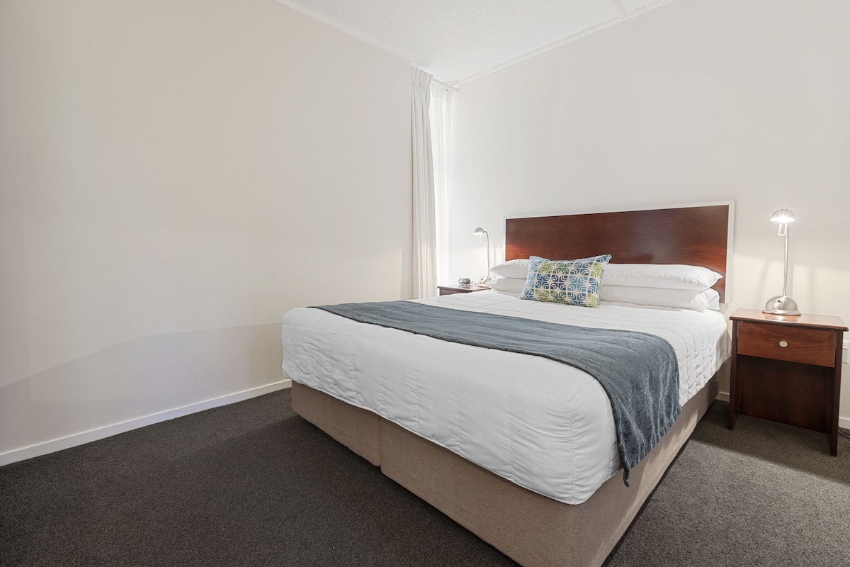 A neatly made bed with a wooden headboard, a bedside table with a lamp, and a window with curtains.
