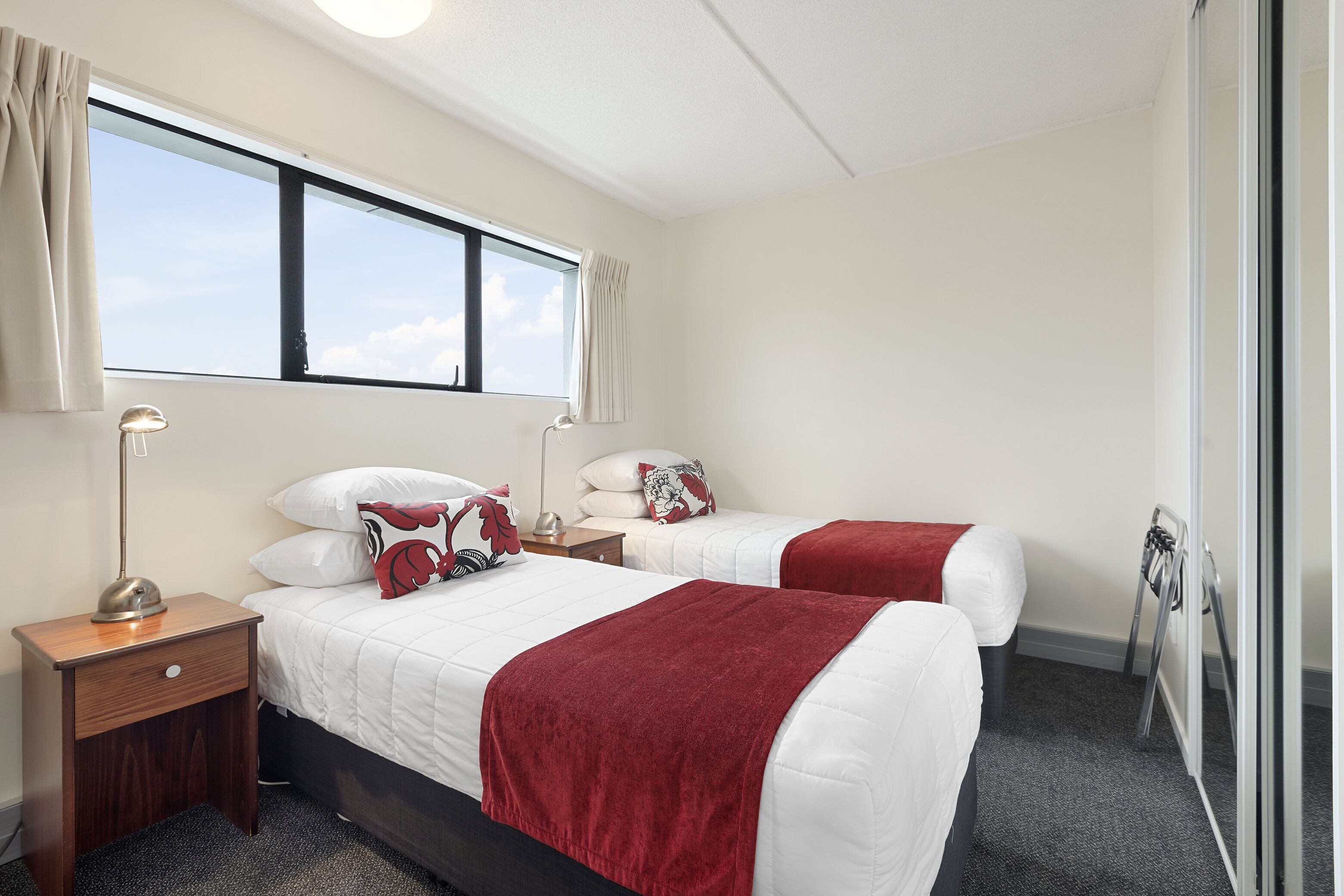 A hotel room with two beds, a nightstand with a lamp, a window with curtains, and a door with a mirror.