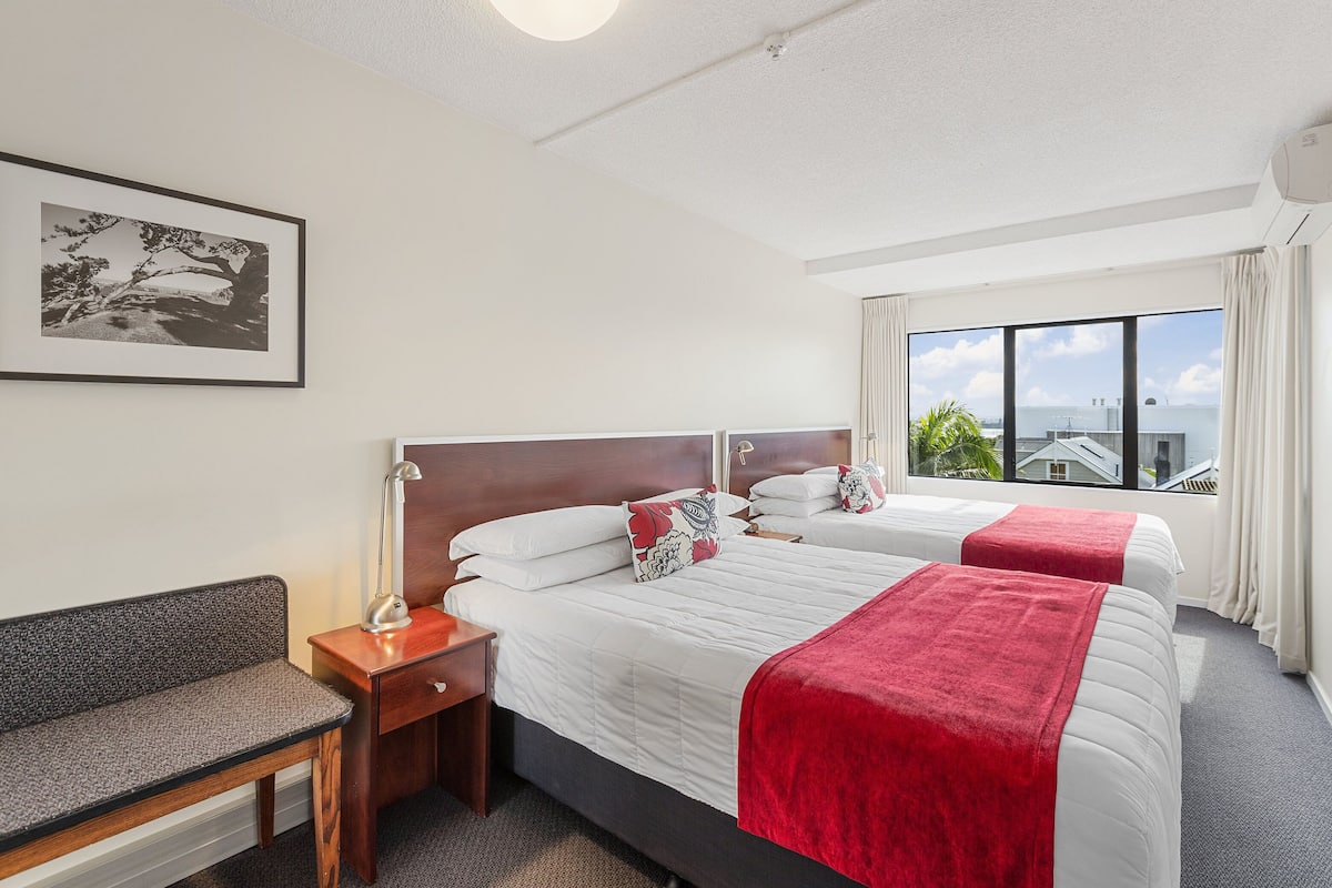 A hotel room with two beds, a bench, a desk, and a large window with a view.