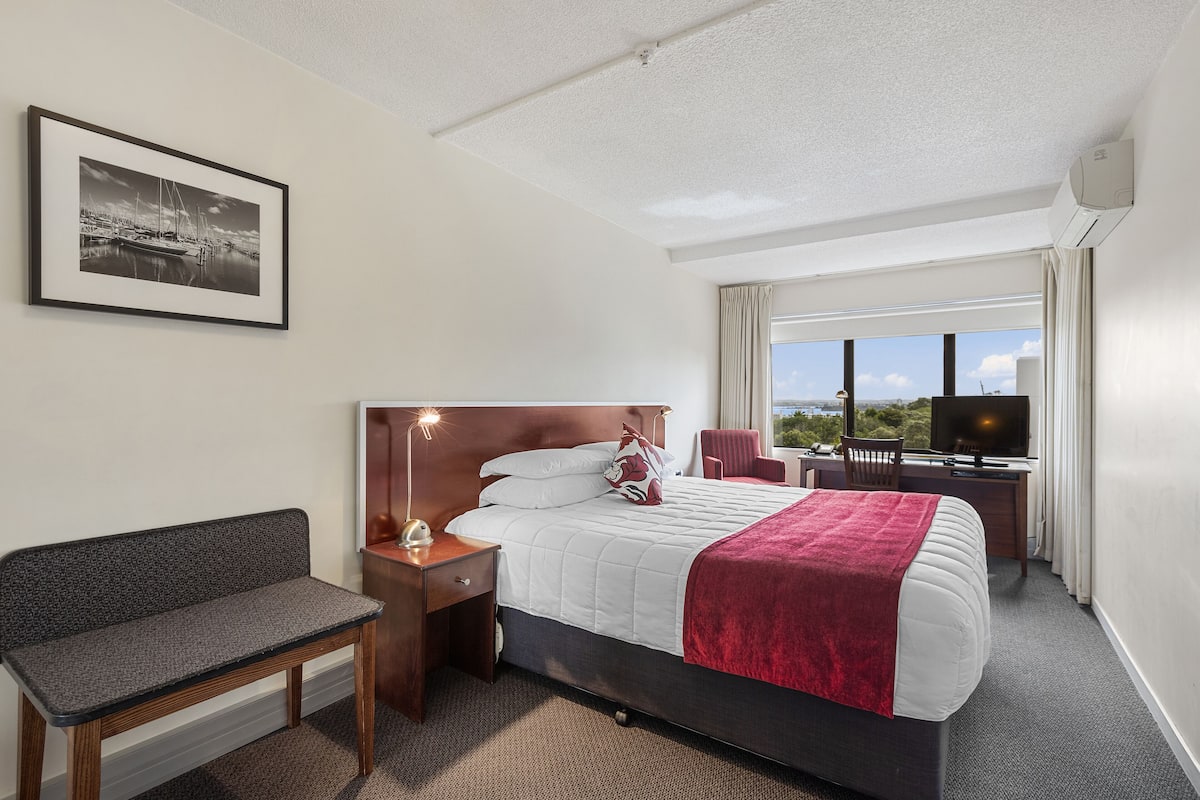 A hotel room with a bed, a desk, a chair, a bench, a TV, and a window with a view.