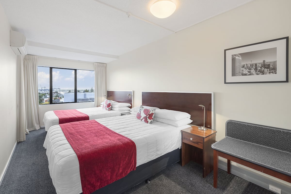 A hotel room with two beds, a desk, a bench, a framed cityscape picture, and a view of the city through large windows.