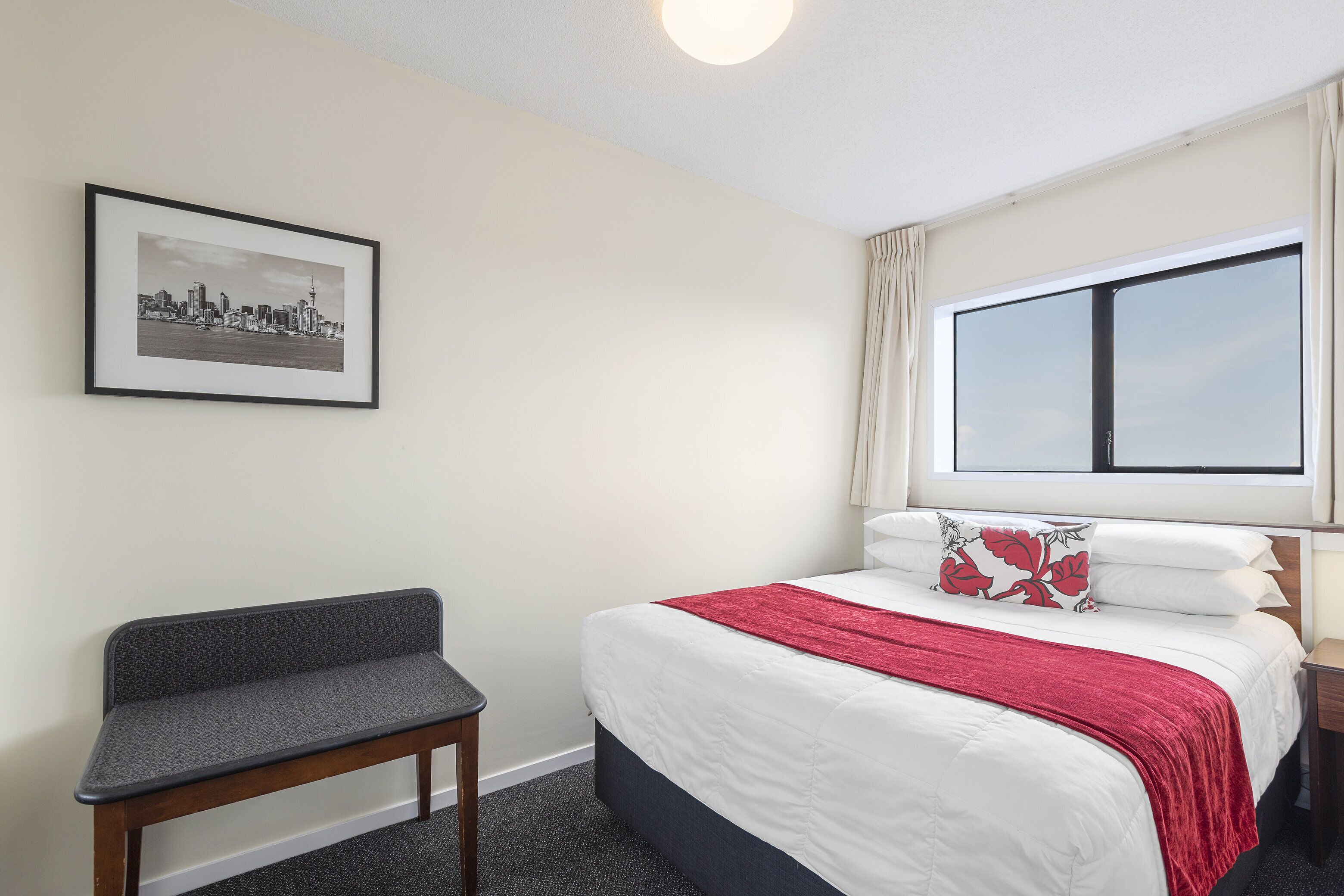 A hotel room with a bed, a bench, a framed cityscape picture, and a large window.