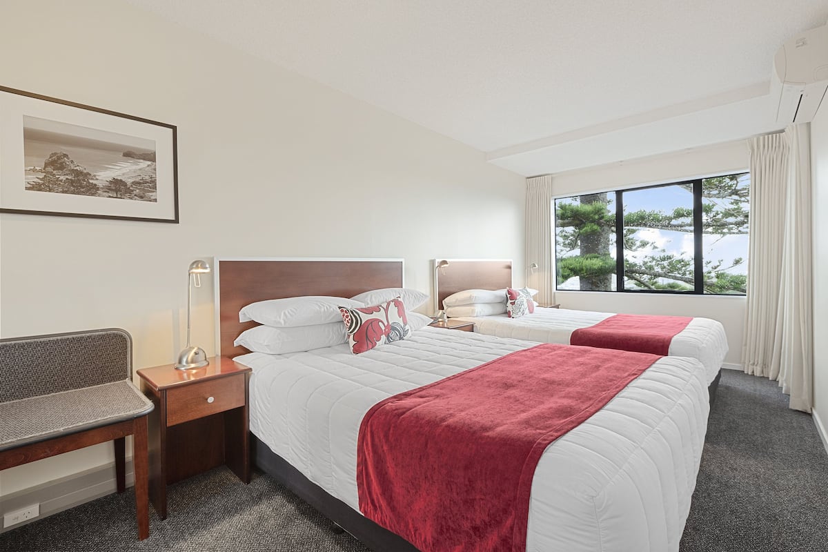 A hotel room with two beds, a desk, a chair, a bedside table, a lamp, and a window with a view of trees.