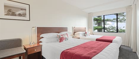 A hotel room with two beds, a desk, a chair, a bedside table, a lamp, and a window with a view of trees.