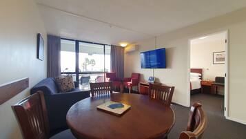 Habitación de hotel con zona de comedor, televisor montado en la pared y vistas a palmeras desde el exterior.