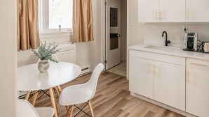 A modern kitchen with white cabinets, a sink, and a coffee maker. There is a dining area with a white table and chairs. - Aurora Inn (Kingston)