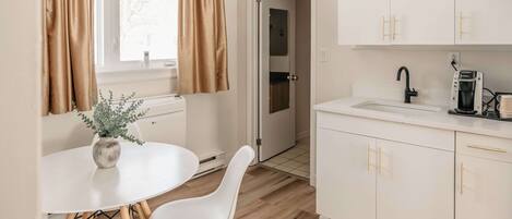 Cocina moderna con armarios blancos, un lavamanos y una cafetera. Hay un comedor con mesa y sillas blancas.