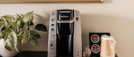 Een Keurig koffiezetapparaat op een tafel, met een plant en een lijst met een schilderij op de achtergrond.