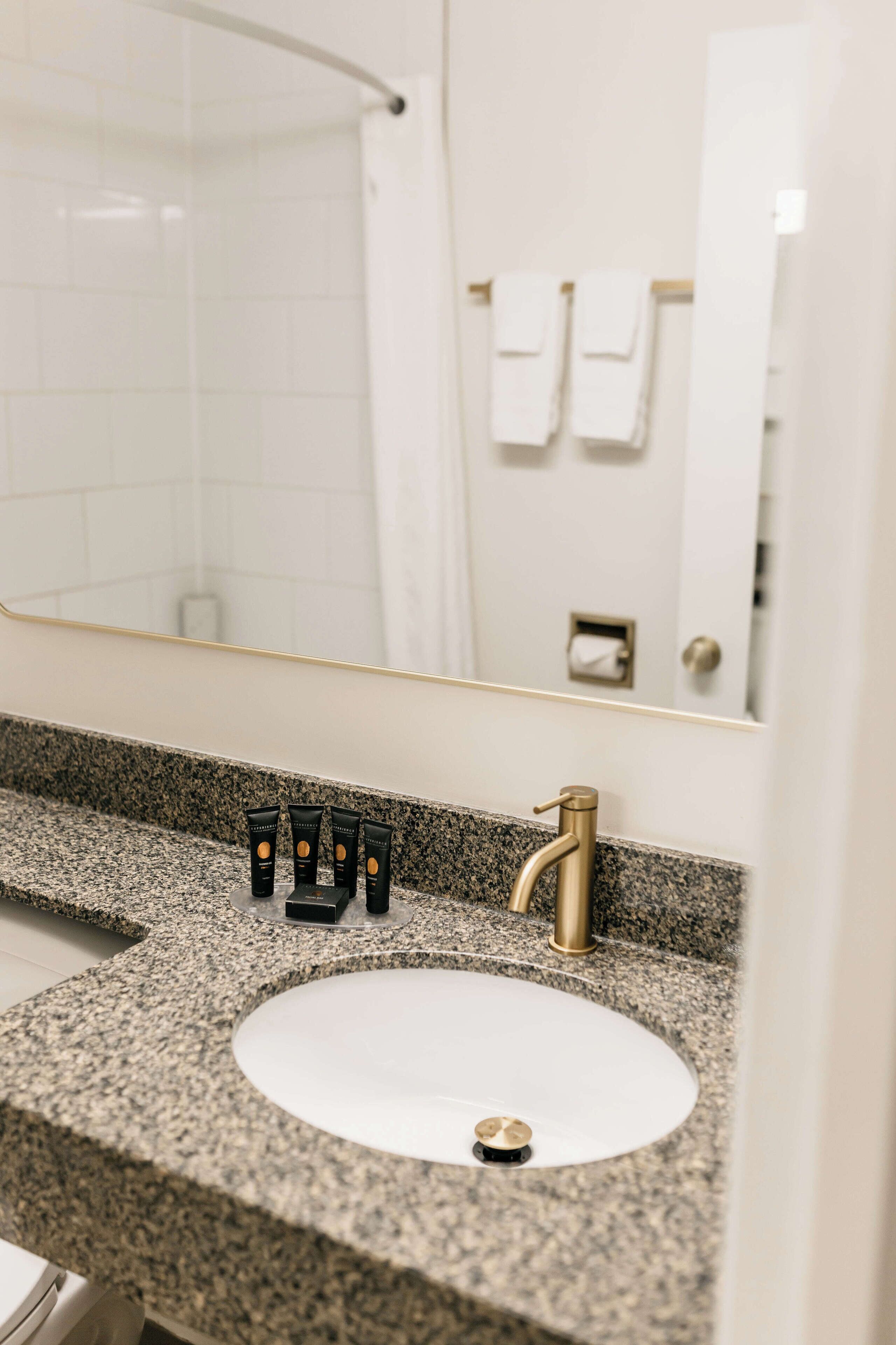 A bathroom with a granite countertop, a white sink, and a gold faucet. There are three black tubes on the counter.