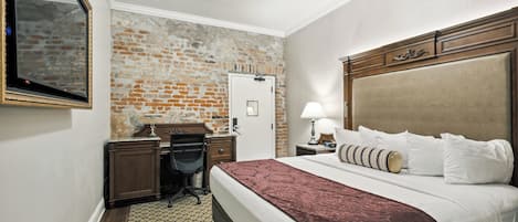 A hotel room with a brick accent wall, a large bed with a headboard, a desk with a chair, a lamp, and a mirror.
