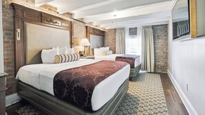 A hotel room with a brick accent wall, two beds with patterned bedding, a large mirror, and a patterned rug. - French Market Inn (New Orleans)