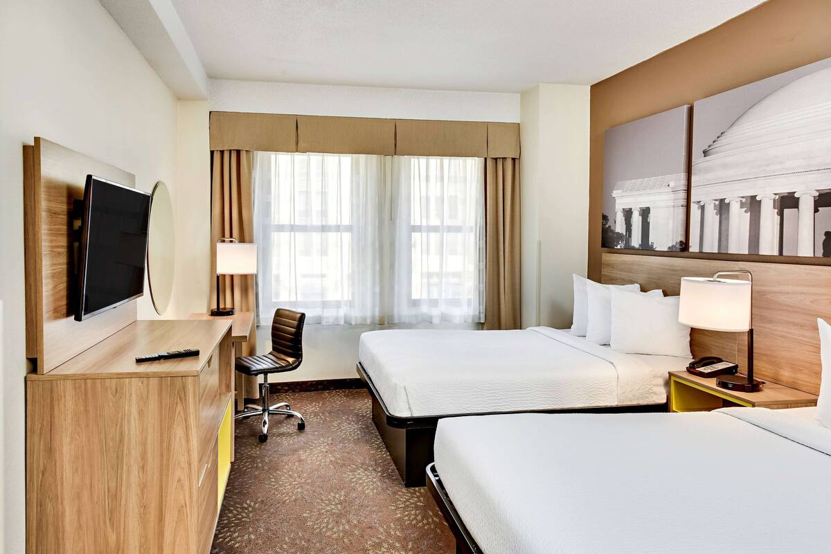 Habitación de hotel con dos camas, un escritorio con televisor, una silla, una lámpara y un cuadro grande en la pared.