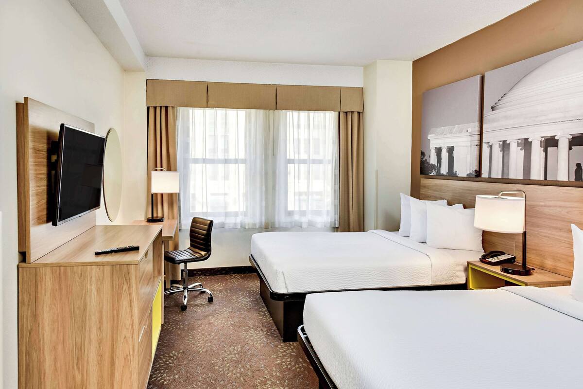 Habitación de hotel con dos camas, un escritorio con televisor, una silla y un ventanal con cortinas.