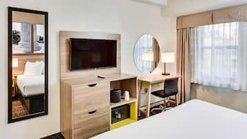 A hotel room with a bed, a desk with a chair, a television, a mirror, and a window with curtains.