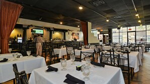 Um restaurante com várias mesas preparadas para refeições, um espaço de bar e uma tela de televisão.