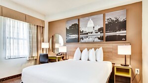 Quarto de hotel com uma cama grande, duas mesas de cabeceira com abajures, uma escrivaninha com cadeira, um espelho grande e três quadros na parede.