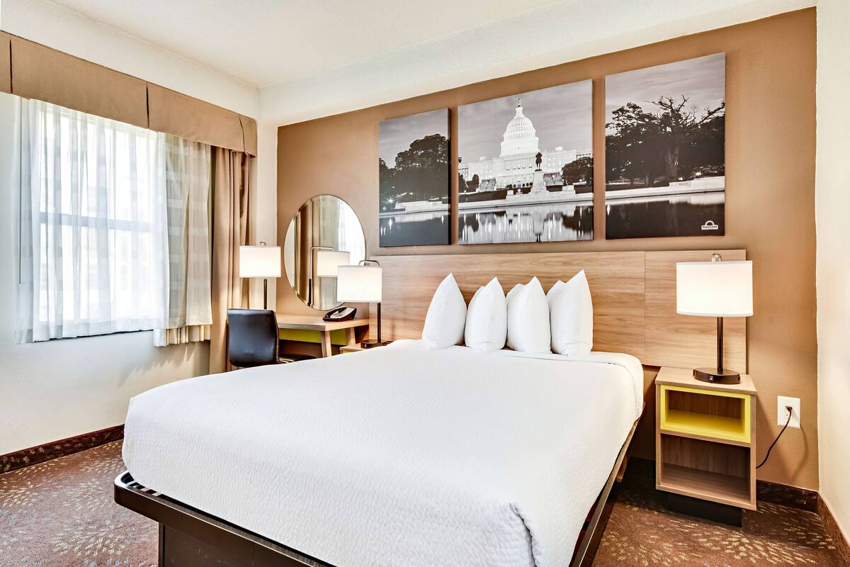 Habitación de hotel con una cama grande, dos mesitas de noche con lámparas, un escritorio con silla, un espejo amplio y tres cuadros enmarcados en la pared.