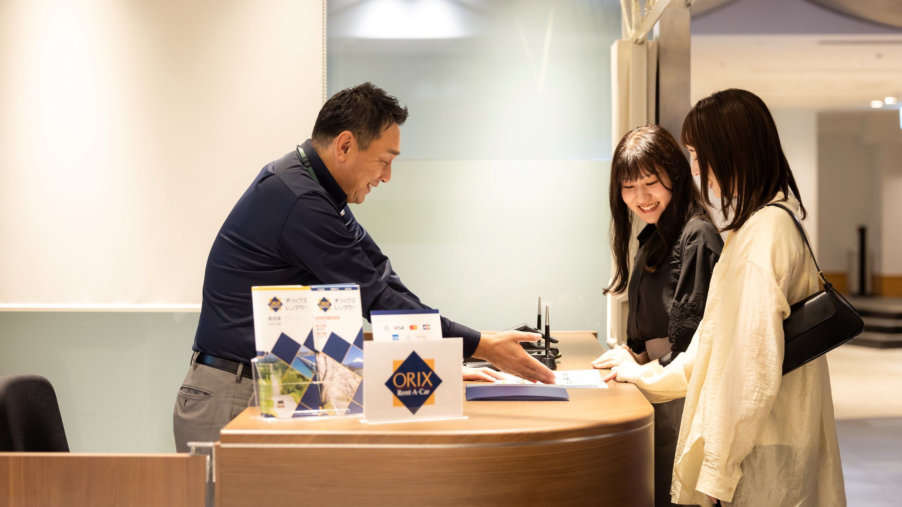 A reception desk with a sign for ORIX Rent-A-Car, a man and two women interacting, and a Visa card acceptance.