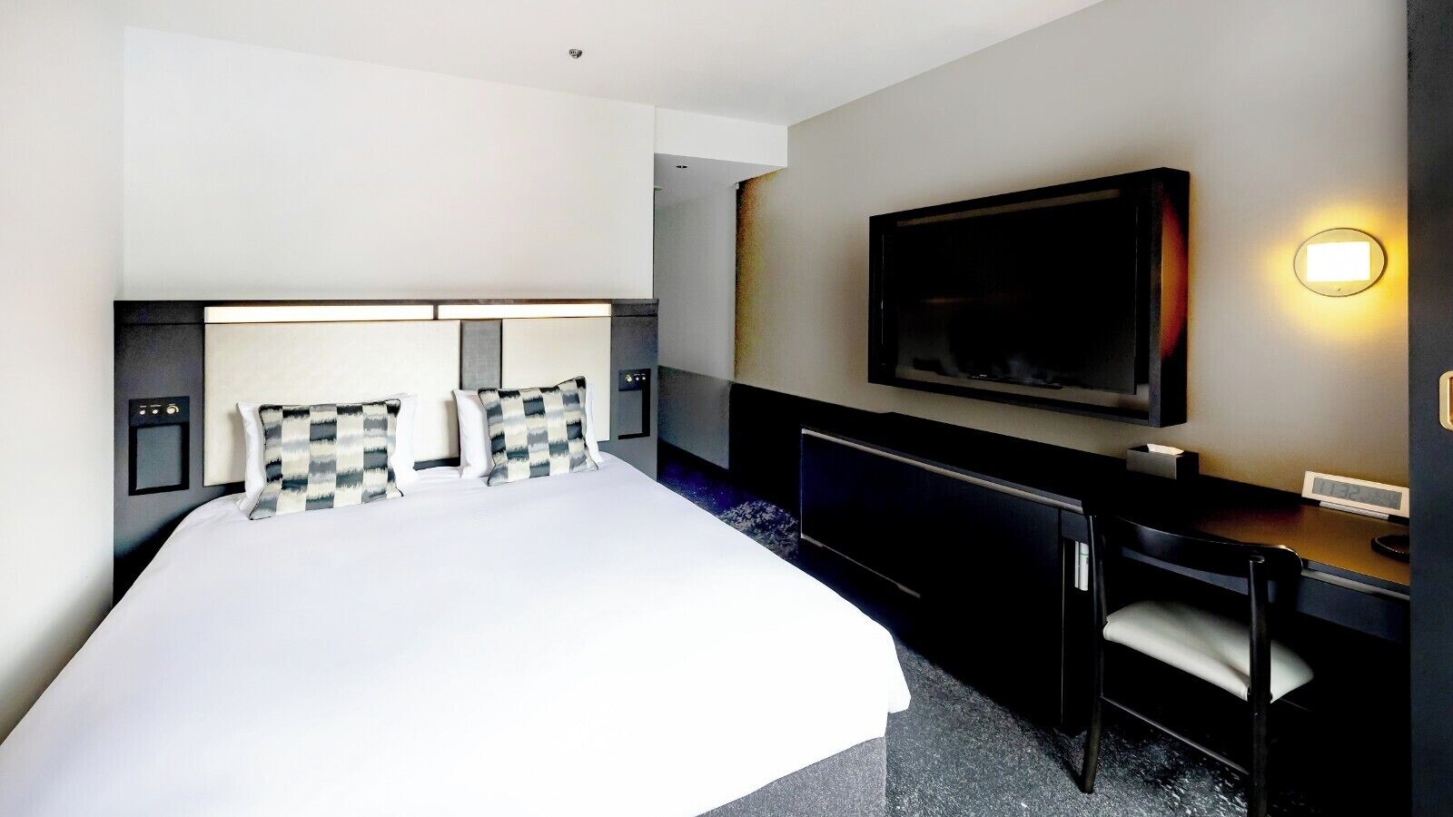 A hotel room with a large bed, a desk, a chair, a flat-screen TV, and a wall-mounted light.