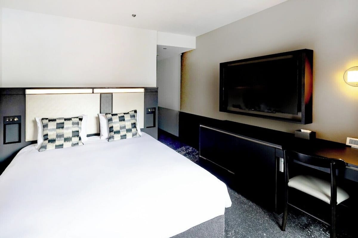 A hotel room with a large bed, a desk, a chair, a flat-screen TV, and a wall-mounted light.