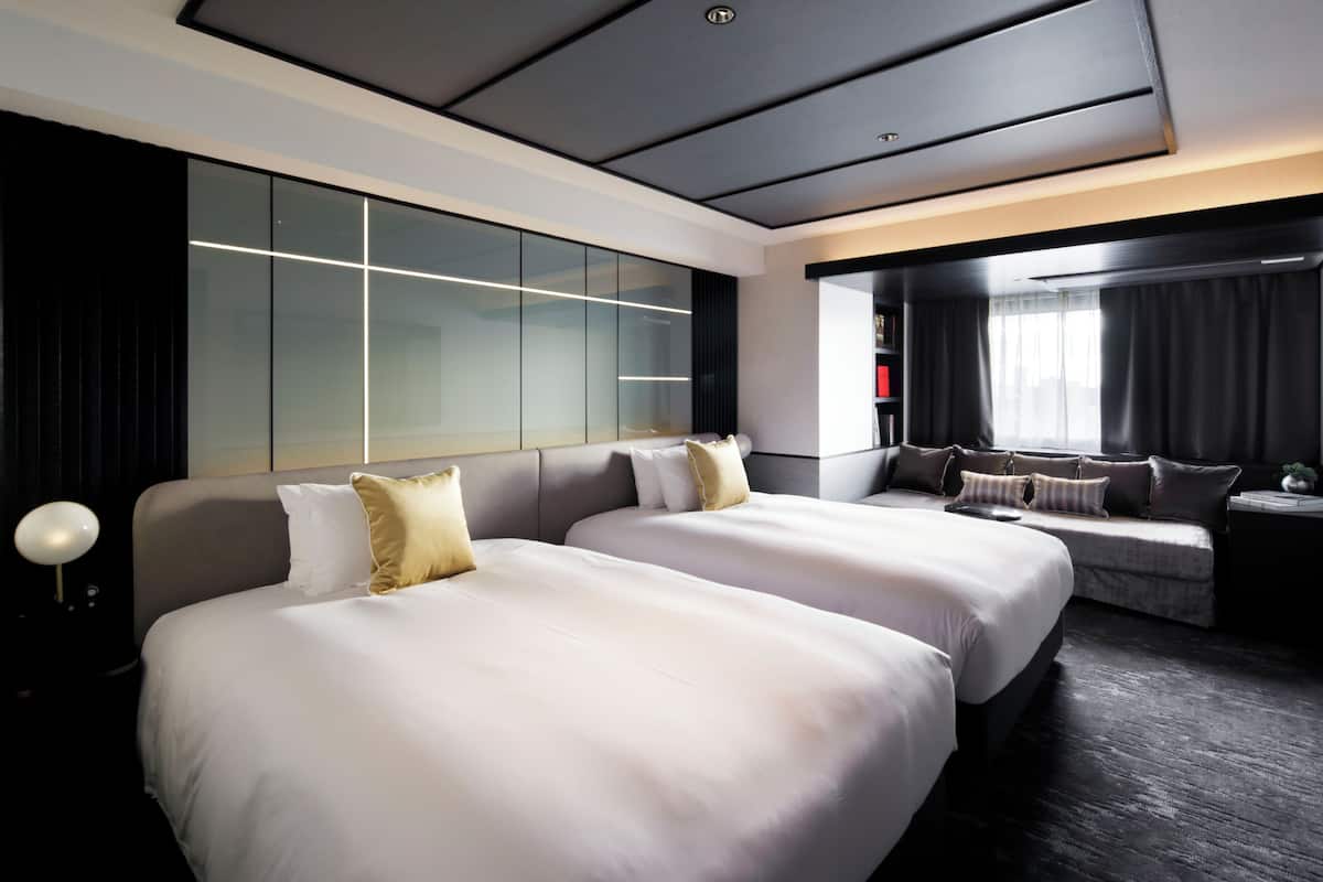 A modern hotel room with two large beds, a seating area, a large window, and a modern ceiling design.
