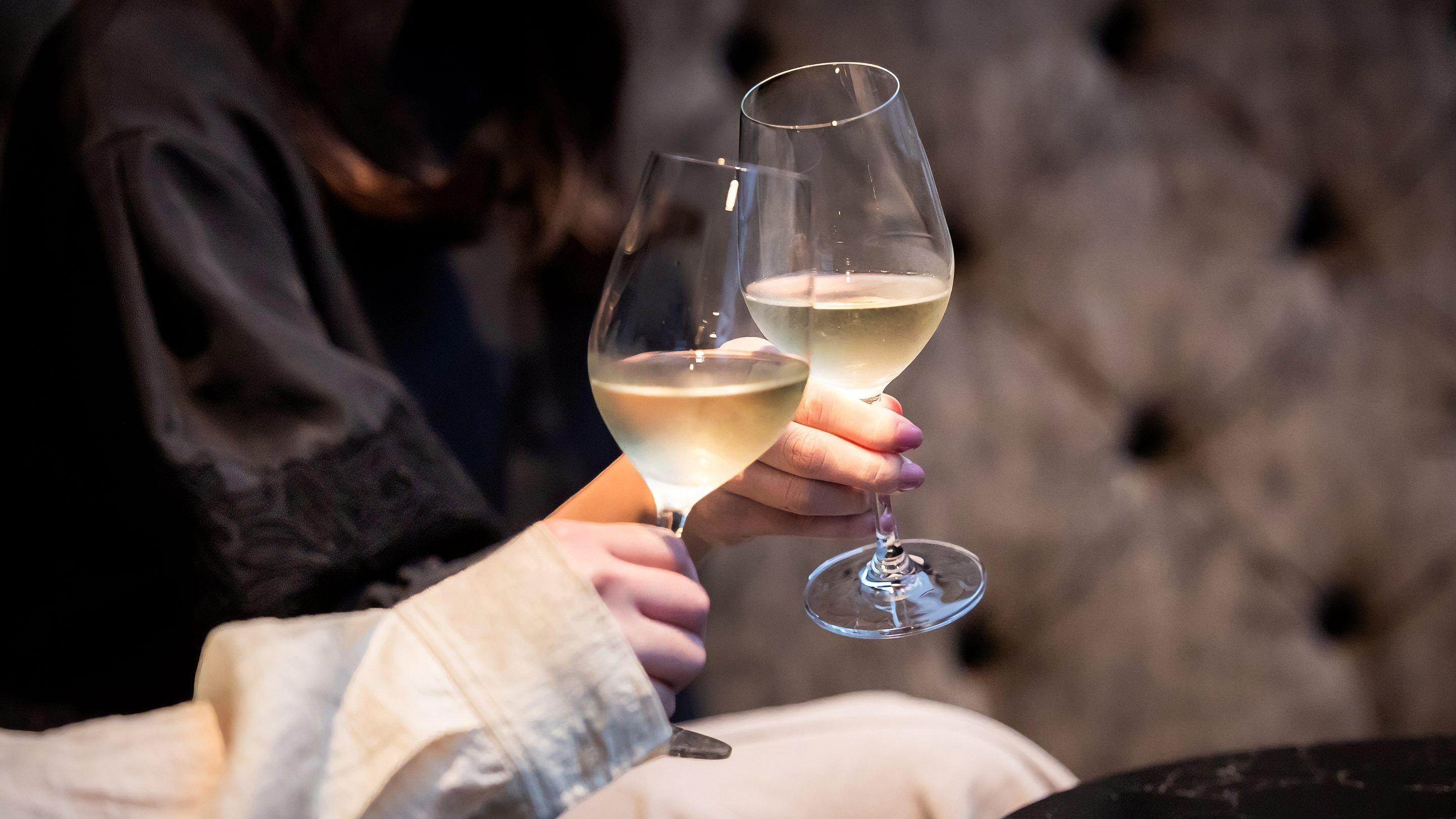 A person holding two wine glasses, one with a white liquid, against a blurred background.