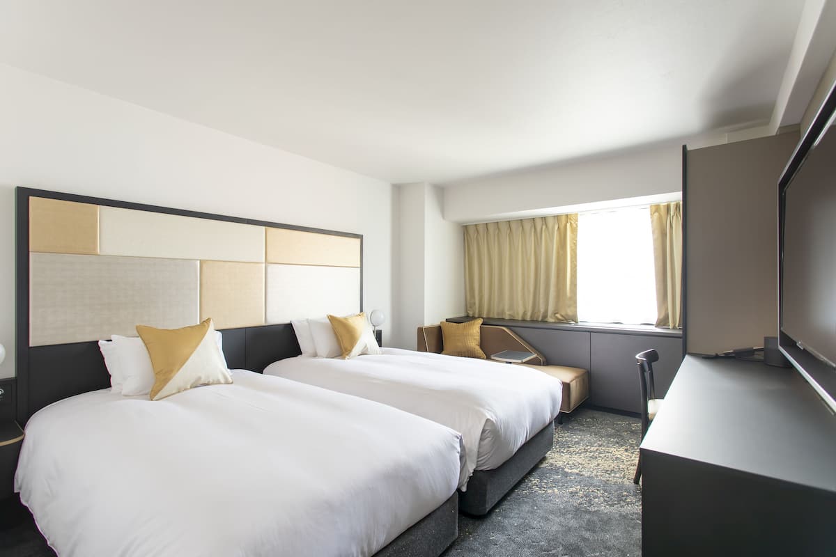 A hotel room with two beds, a desk, a TV, and a window with curtains.