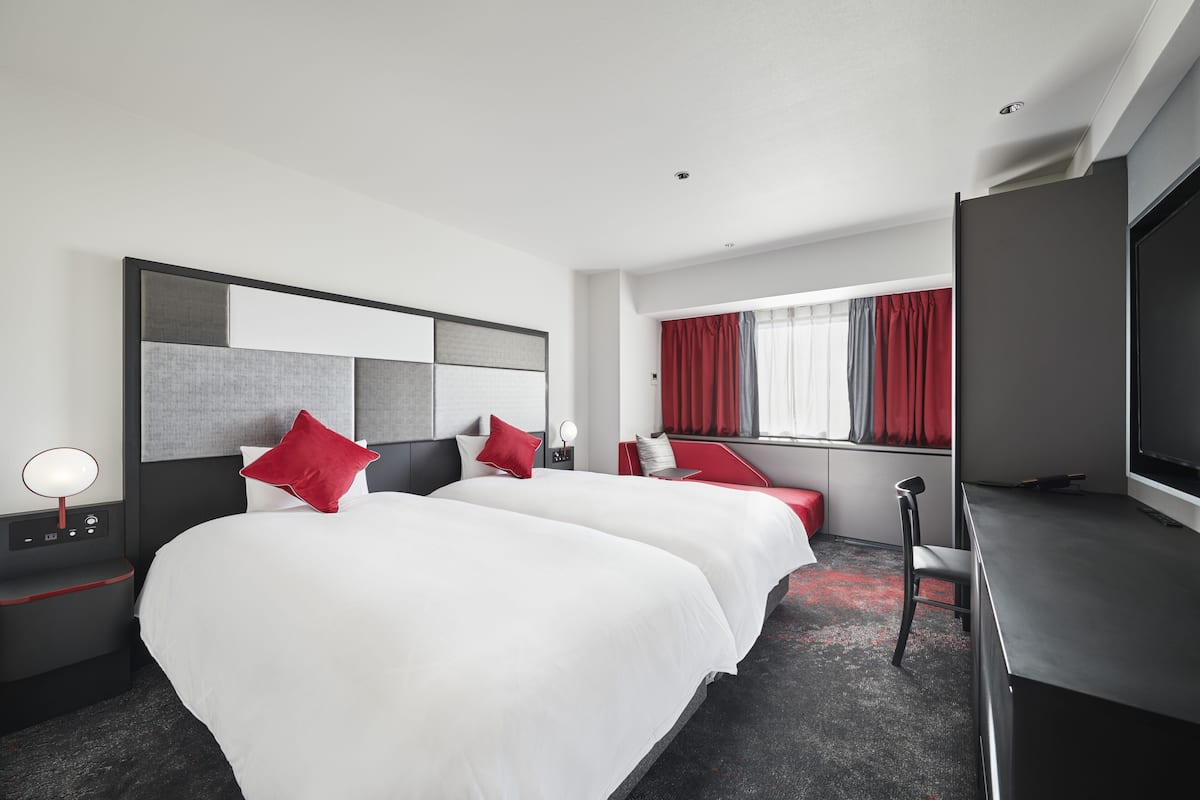A hotel room with two beds, a TV, a desk, and a window with red curtains.