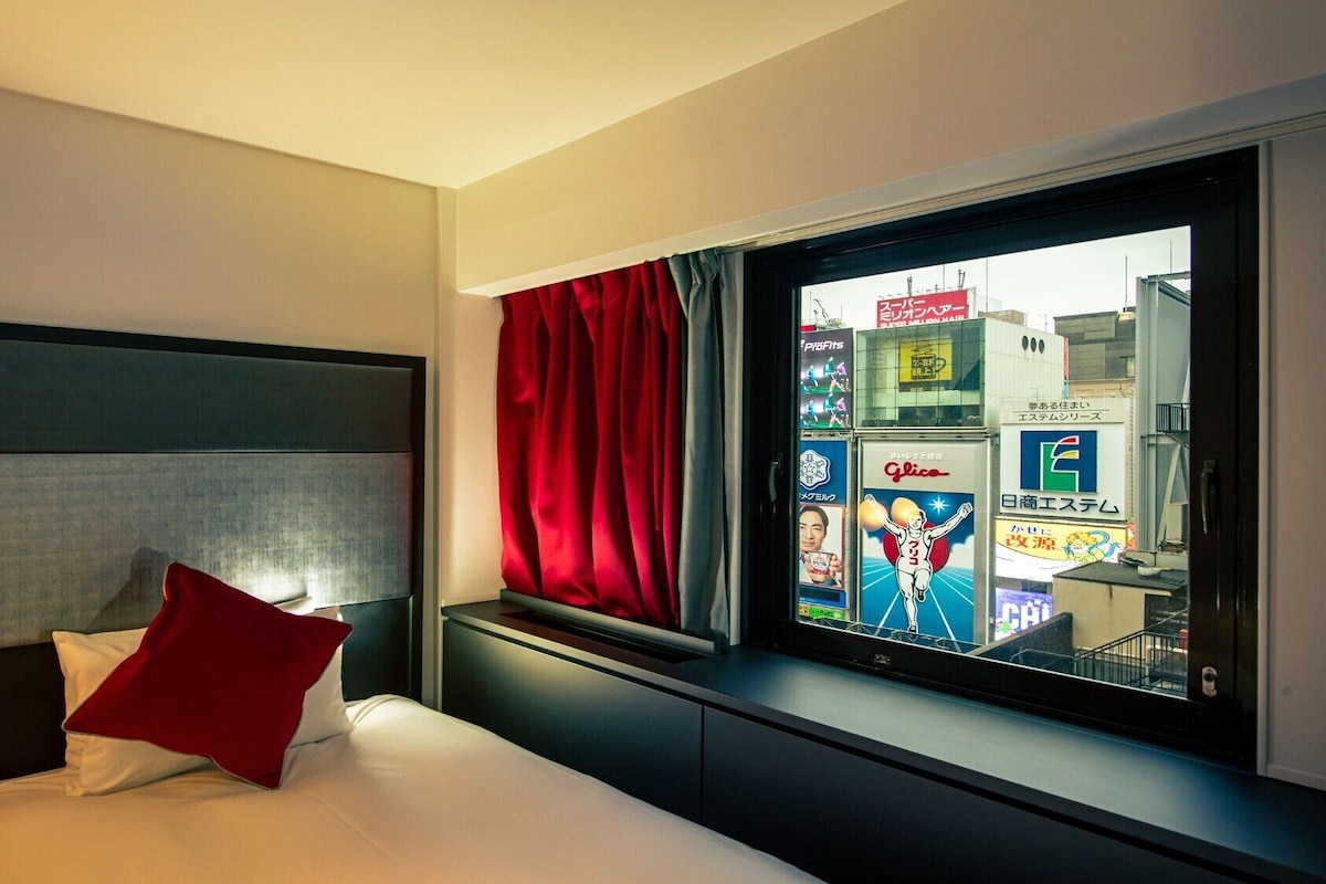 A hotel room with a bed, a nightstand, a lamp, a window with red curtains, and a view of the cityscape.