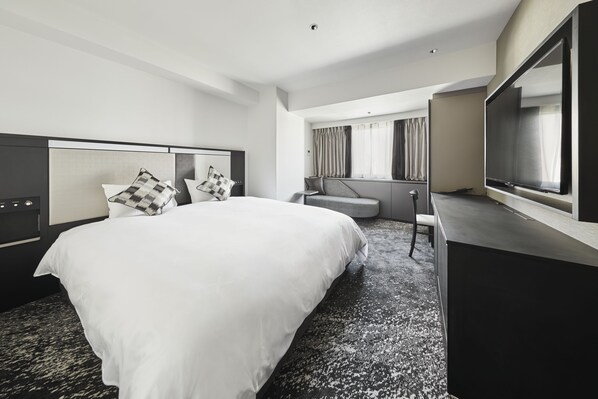 A modern hotel room with a large bed, a desk, a TV, and a seating area with curtains.