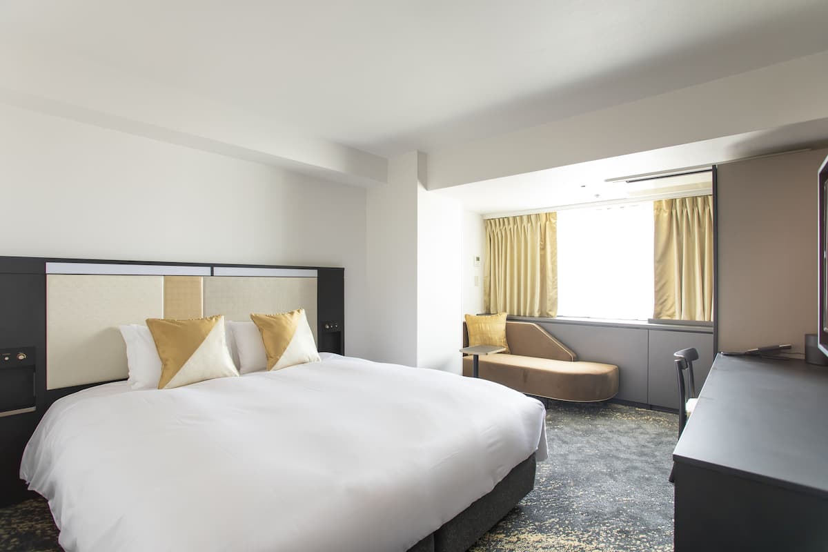 A hotel room with a large bed, a desk, a chair, and a window with curtains.