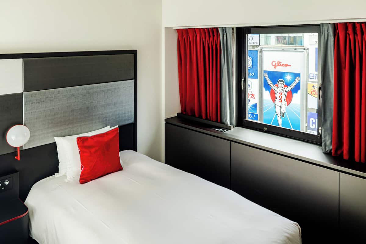 A modern hotel room with a bed, a nightstand, a window with curtains, and a wall-mounted TV displaying an advertisement.