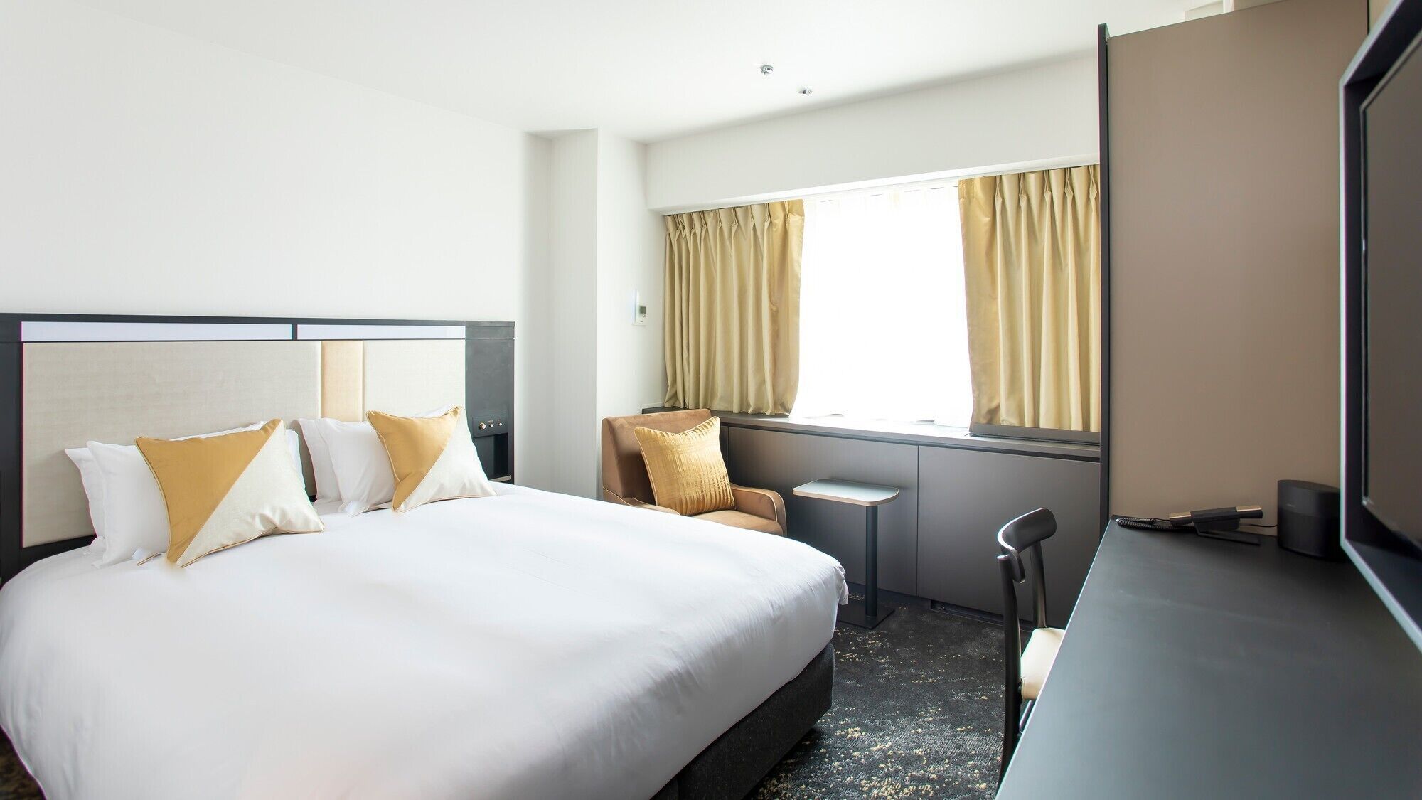 A hotel room with a bed, a chair, a desk, and a large window with curtains.