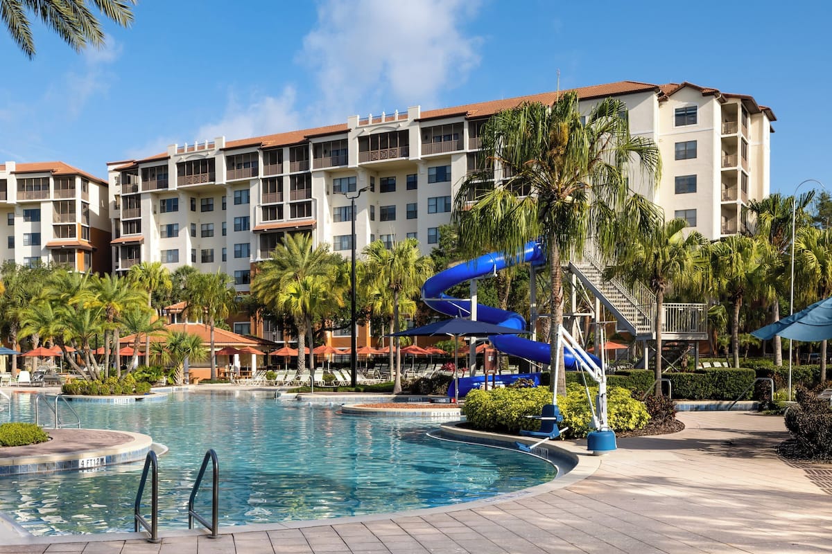 A hotel with a swimming pool, palm trees, and a blue water slide.