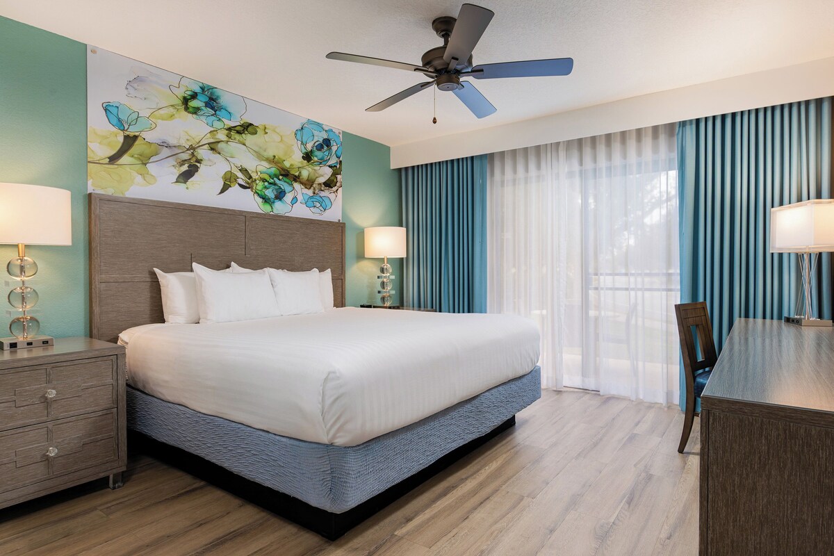 A hotel room with a bed, nightstands, a ceiling fan, a painting, and a view of the outdoors.