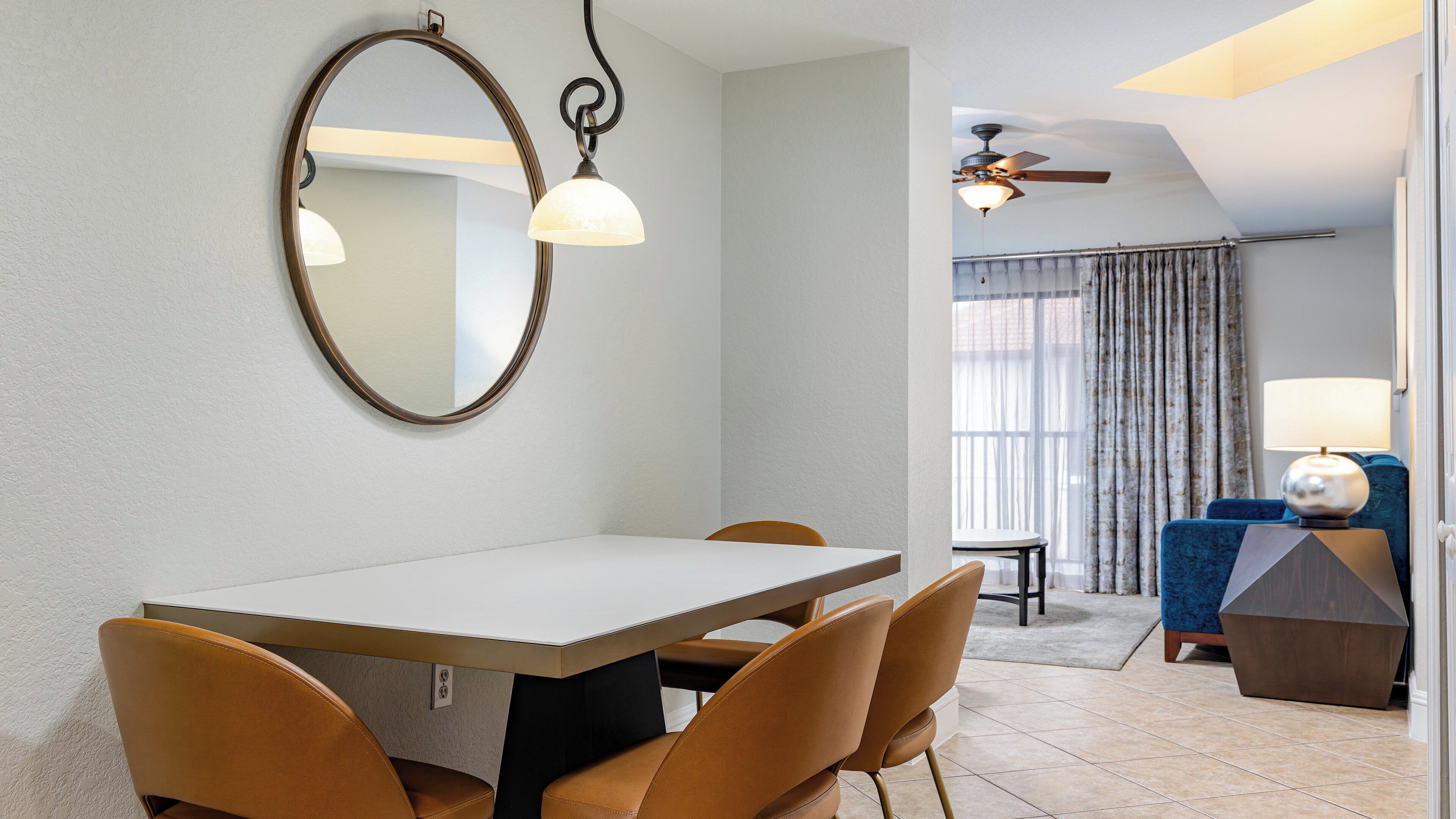 Un área de comedor con una mesa, sillas, un ventilador de techo y vistas a una sala de estar con un sofá y cortinas.