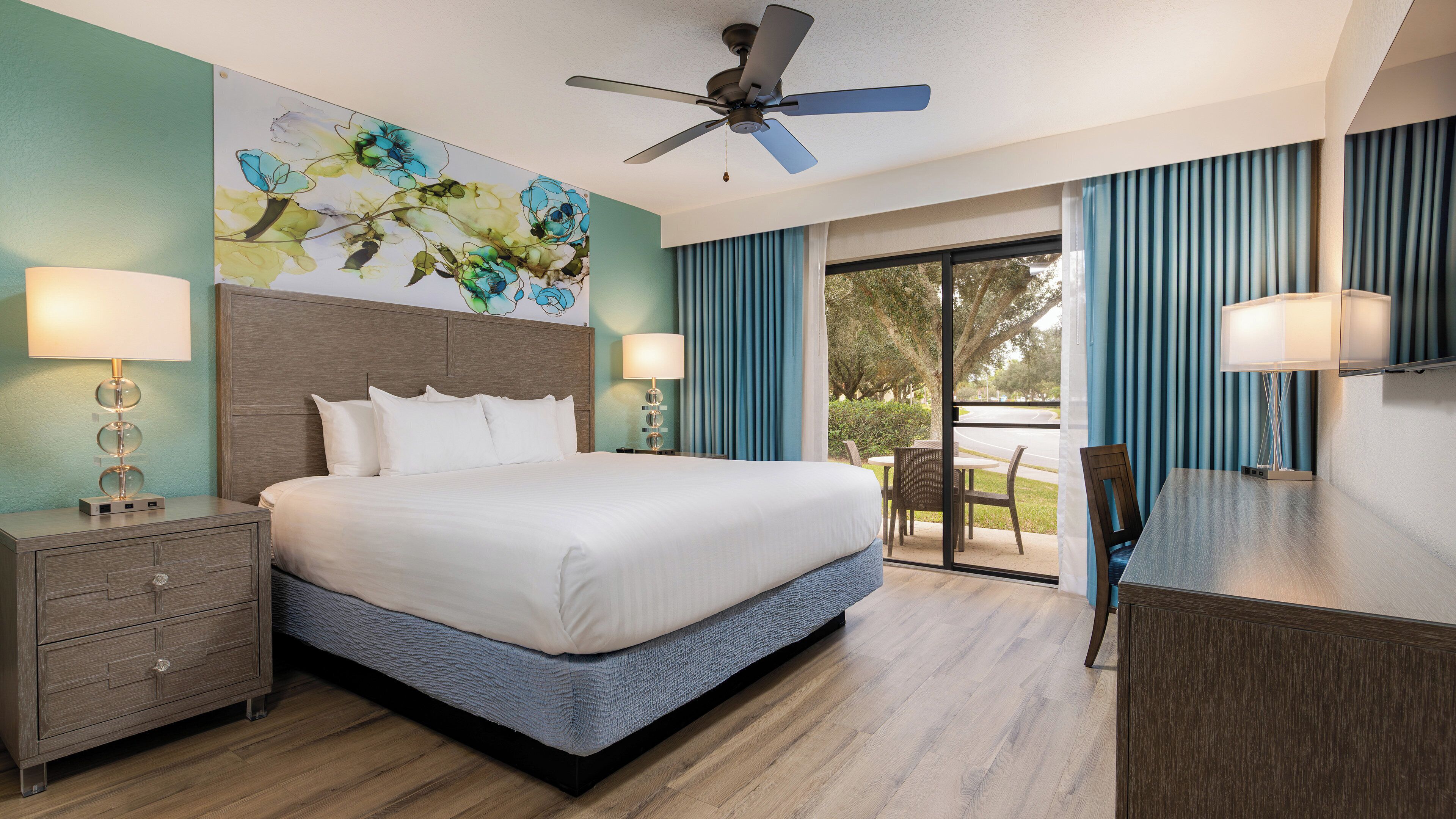 A hotel room with a bed, nightstands, a ceiling fan, a painting, and a view of the outdoors.