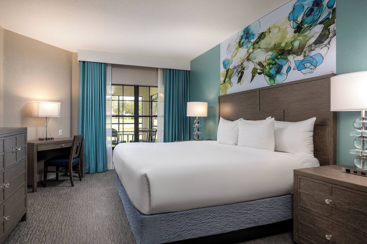 A hotel room with a large bed, a desk with a chair, a television, and a painting on the wall.
