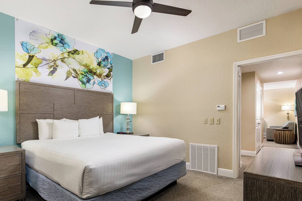 A hotel room with a large bed, two bedside tables with lamps, a ceiling fan, a wall-mounted air conditioning unit, and a door leading to another room.
