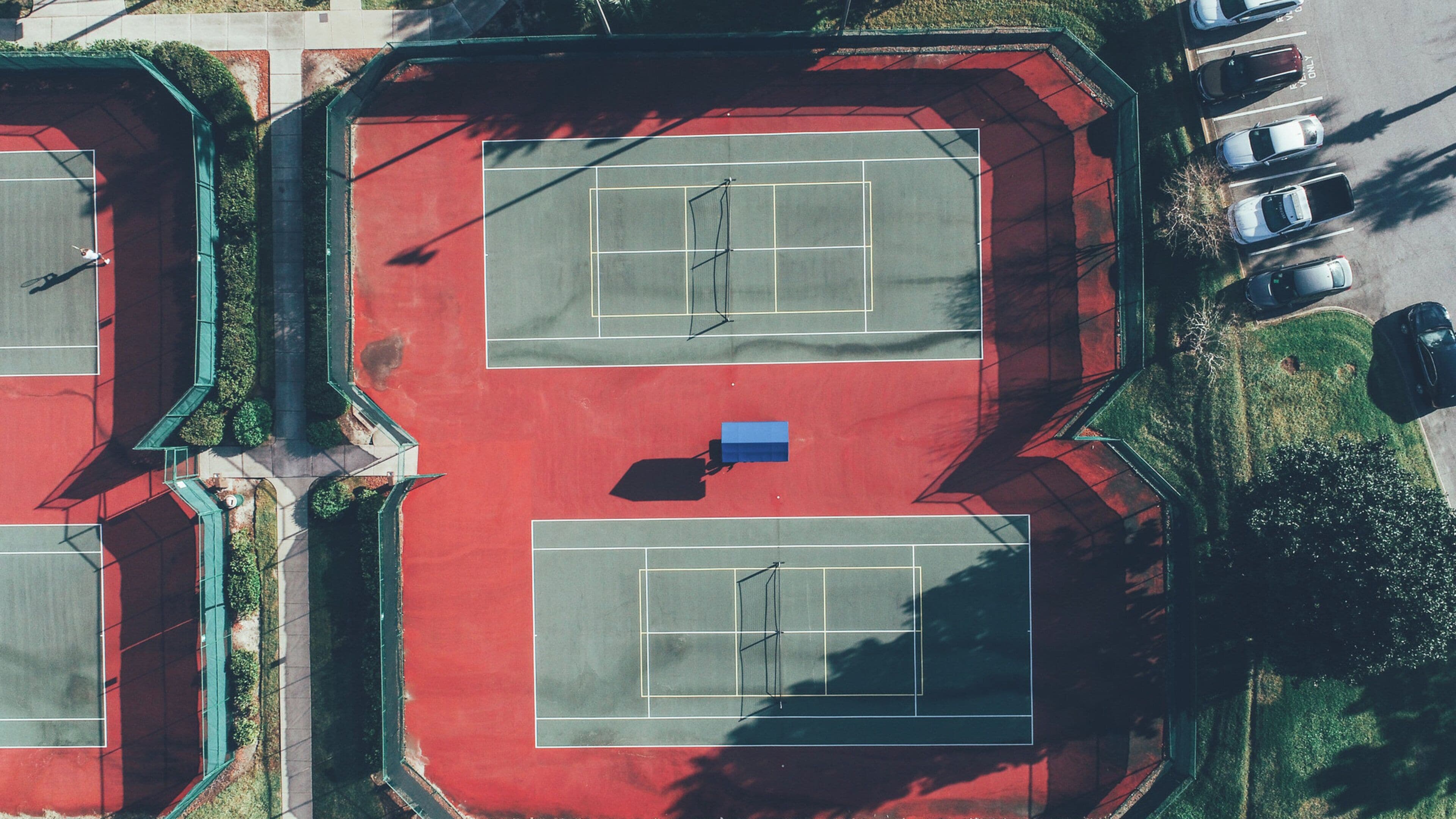 An aerial view of a tennis court complex with multiple courts, a parking area, and surrounding greenery.