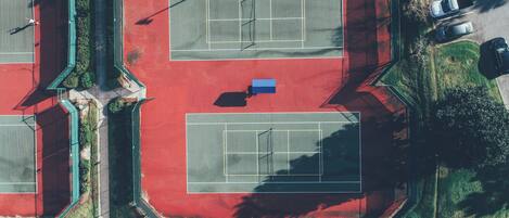 An aerial view of a tennis court complex with multiple courts, a parking area, and surrounding greenery.