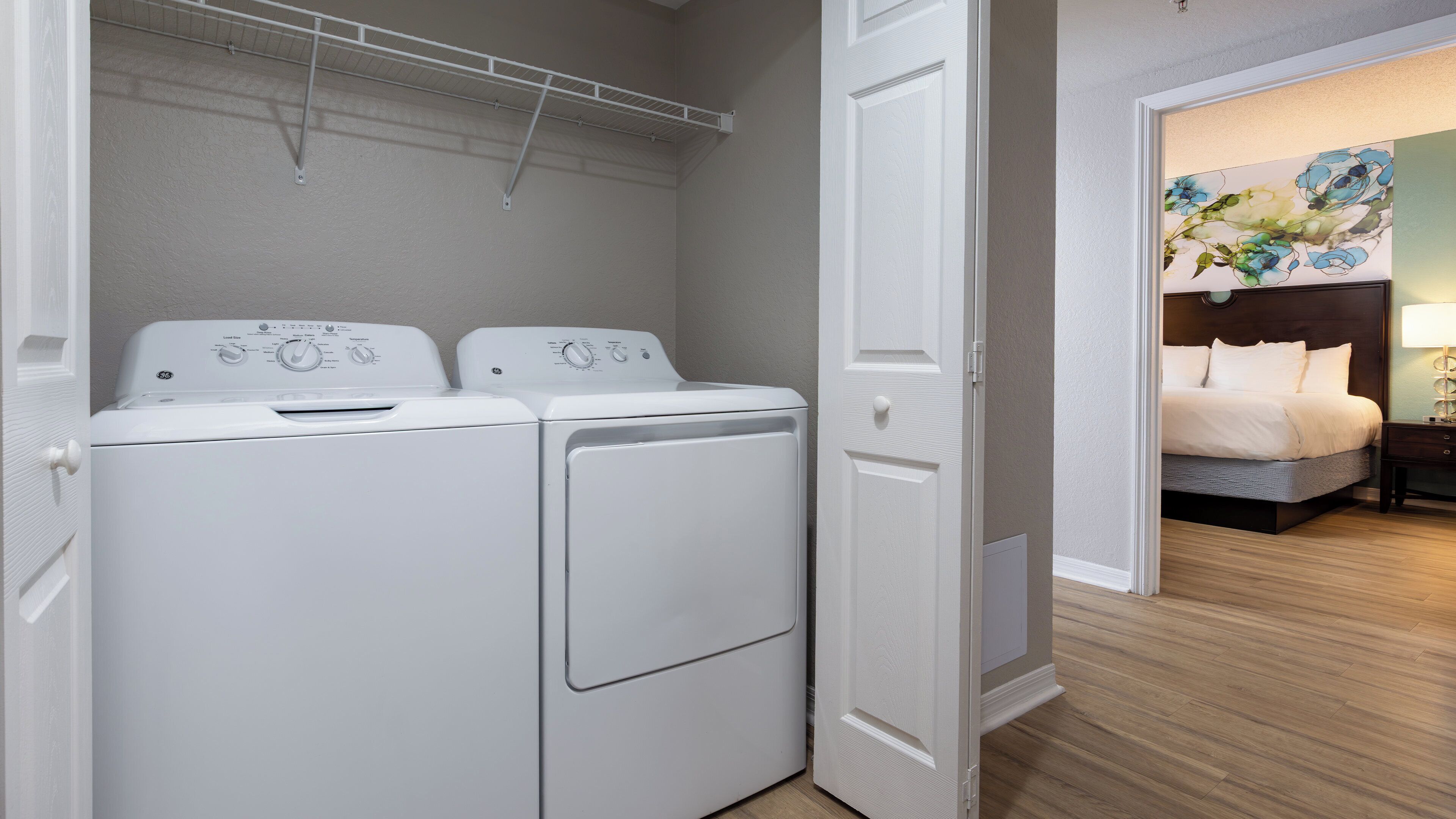 A laundry room with two white front-loading washers and dryers, a white door, and a hallway leading to a bedroom with a bed and a nightstand.