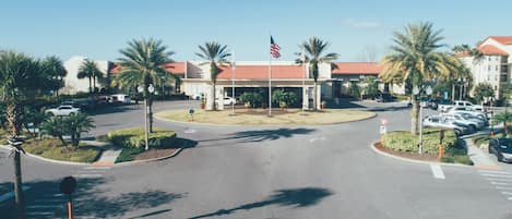 A hotel entrance with a red-roofed building, palm trees, and a parking lot.