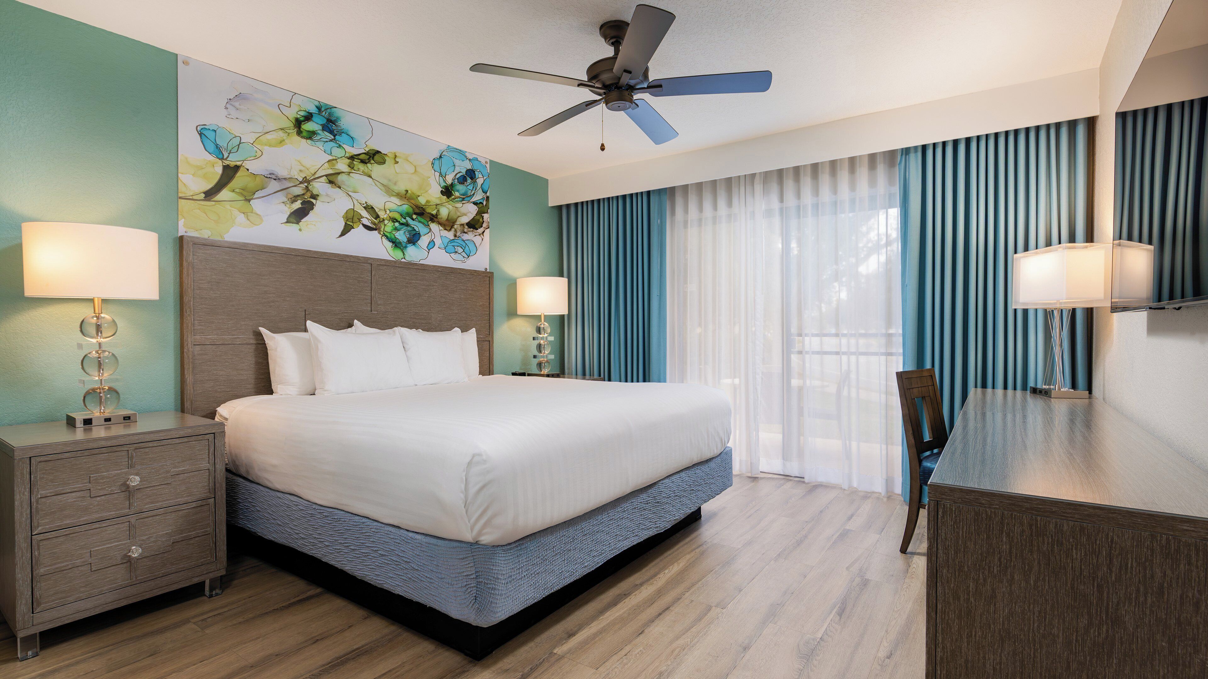 A hotel room with a bed, nightstands, a desk, a chair, a ceiling fan, and a large window with curtains.