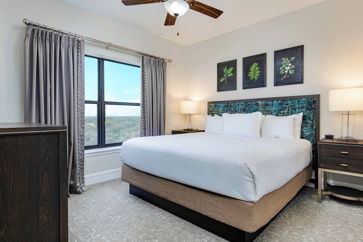 A hotel room with a large bed, a desk, a chair, a window with curtains, a ceiling fan, and artwork on the wall.