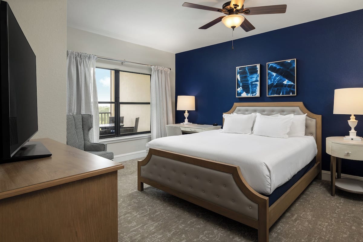 A hotel room with a bed, two bedside tables with lamps, a ceiling fan, a window with curtains, and artwork on the wall.