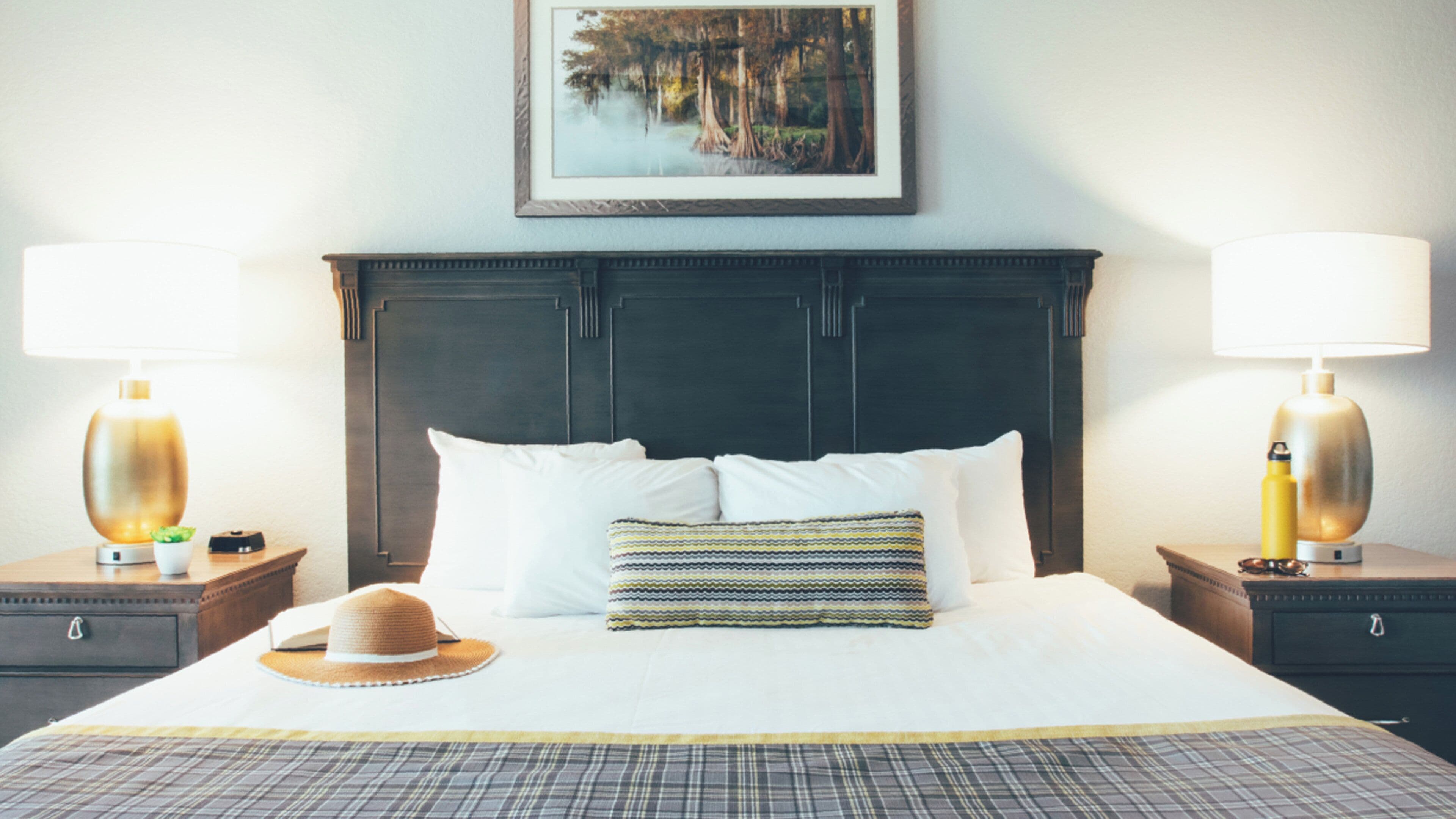 A hotel room with a large bed, two bedside lamps, a framed picture on the wall, and a hat on the bed.