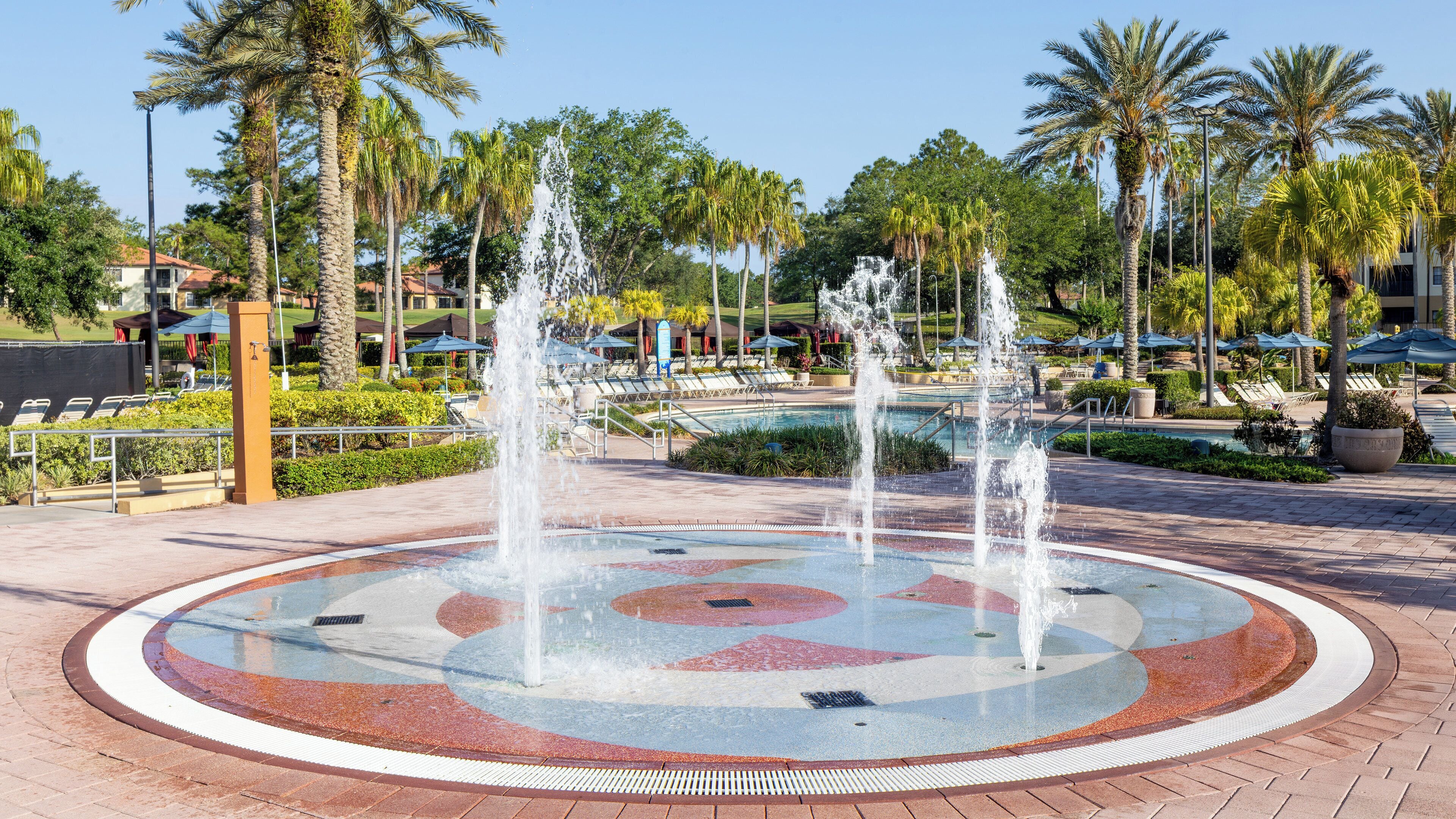 A circular water feature with fountains, surrounded by palm trees and a pool area with lounge chairs.