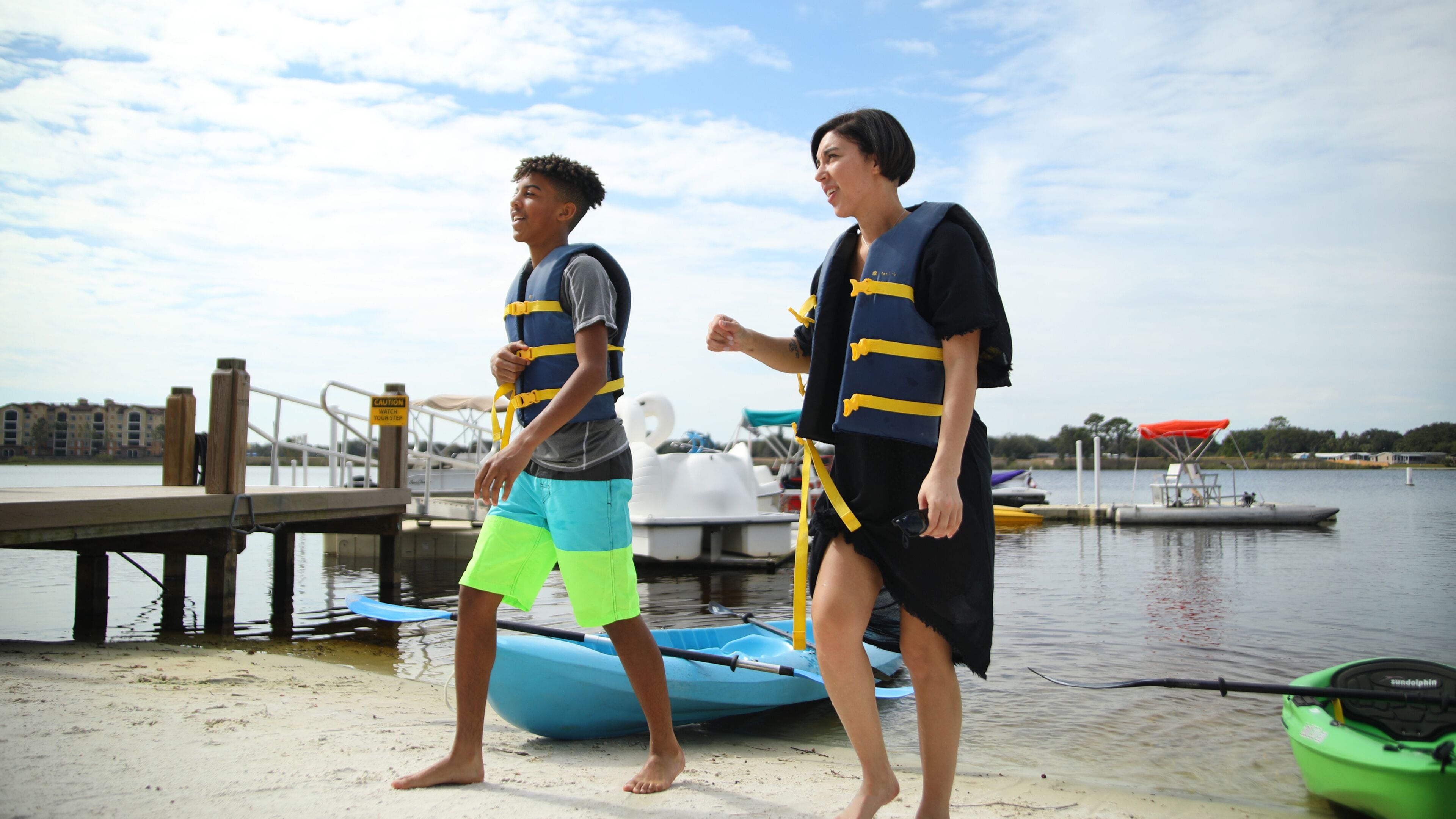 Two people wearing life jackets standing on a dock by a body of water with boats and a building in the background.